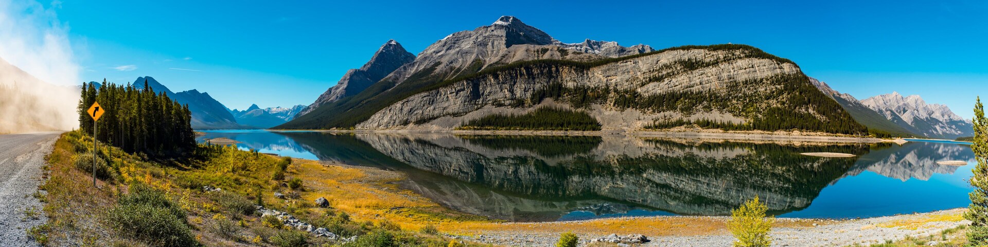 Un lago de montaña con un reflejo claro, una carretera con una señal de curva y una zona boscosa.