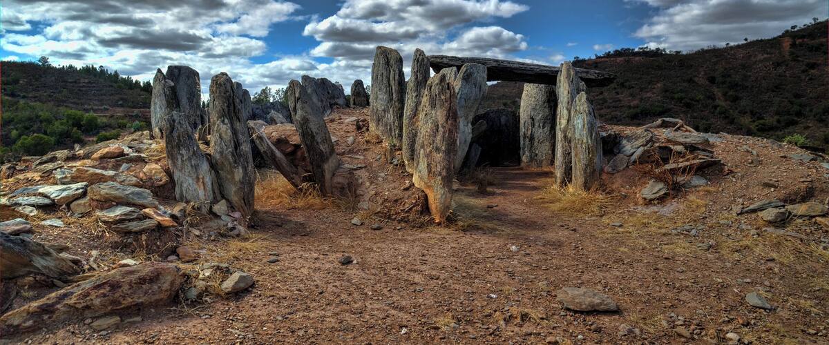 A stone structure with tall pillars in a dry, rocky landscape under a partly cloudy sky.