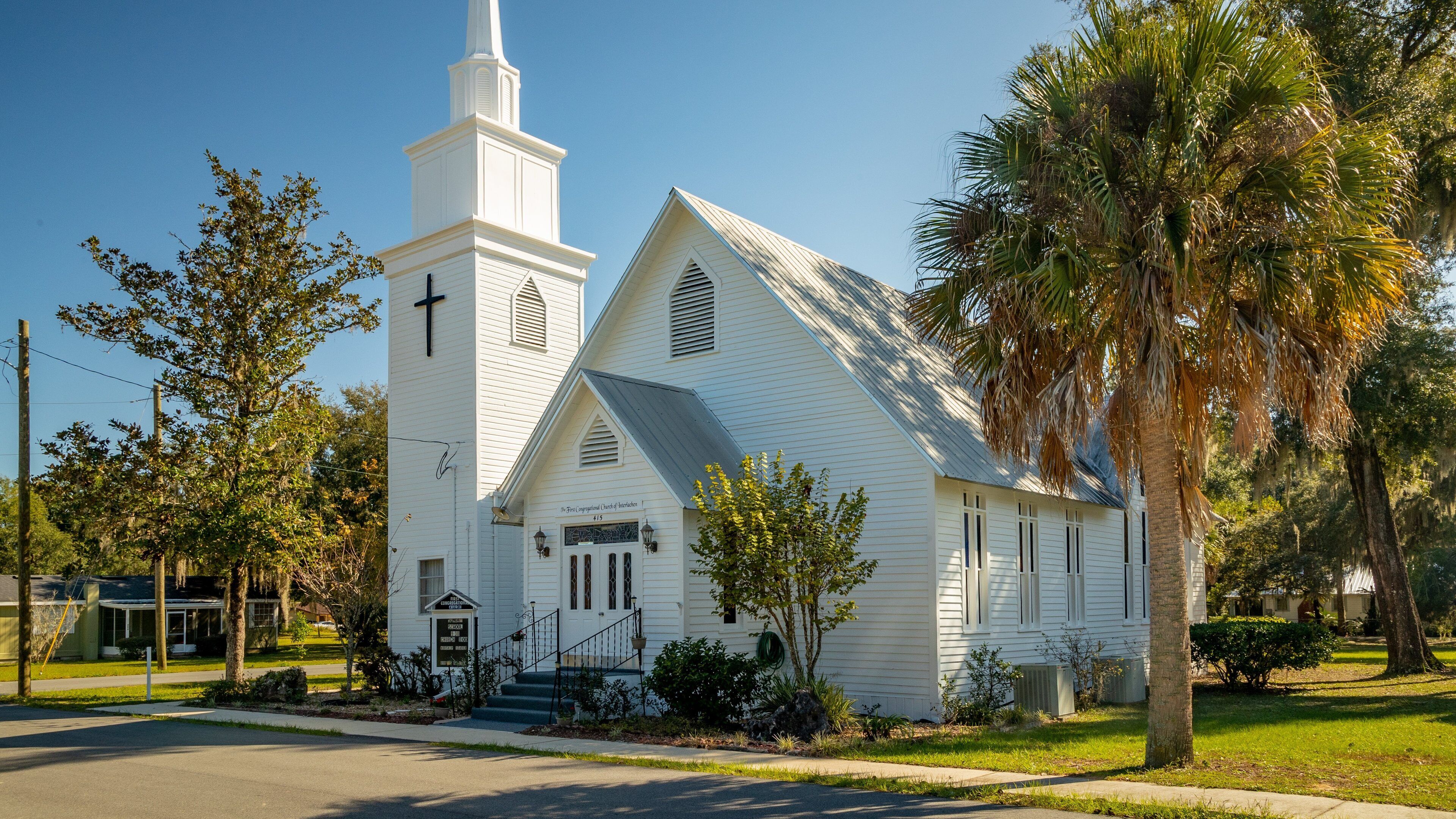 A white church with a steeple and a cross, surrounded by trees and a well-maintained lawn.