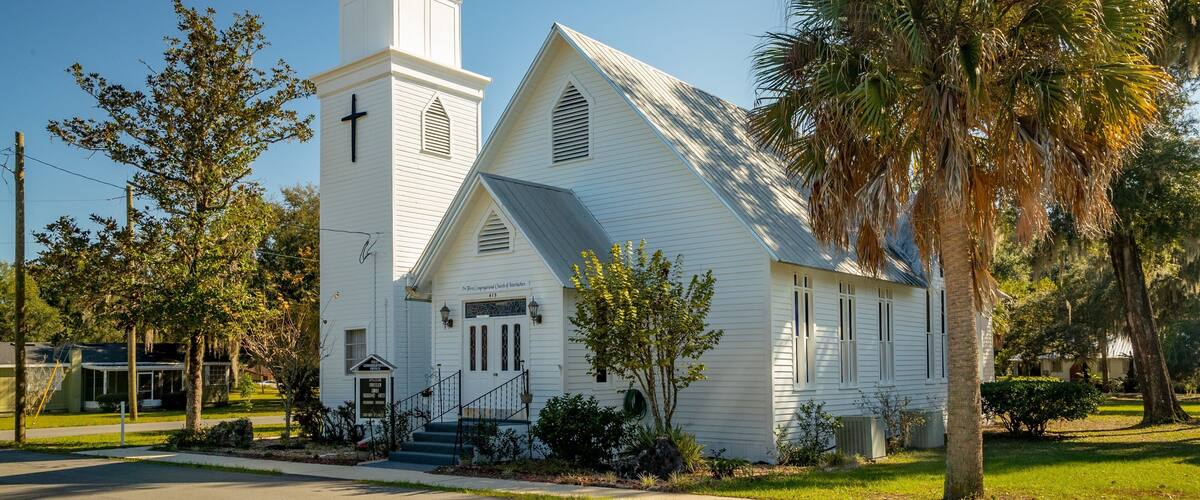 A white church with a steeple and a cross, surrounded by trees and a well-maintained lawn.