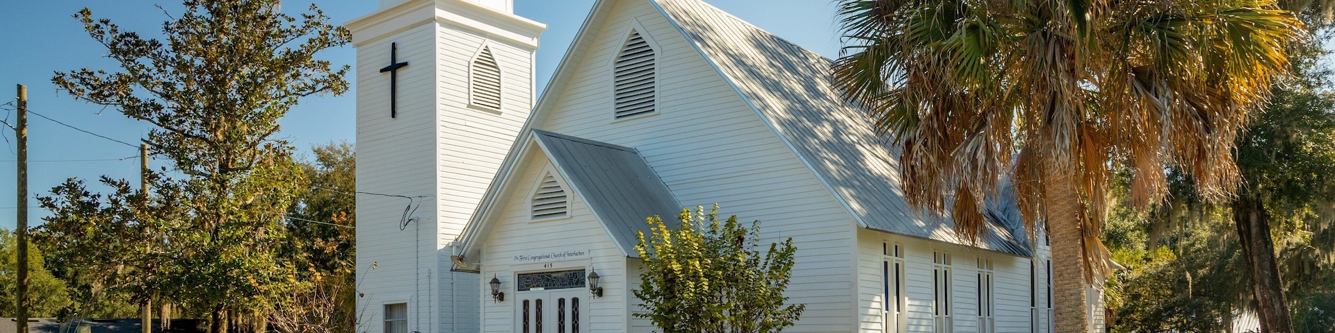 A white church with a steeple and a cross, surrounded by trees and a well-maintained lawn.