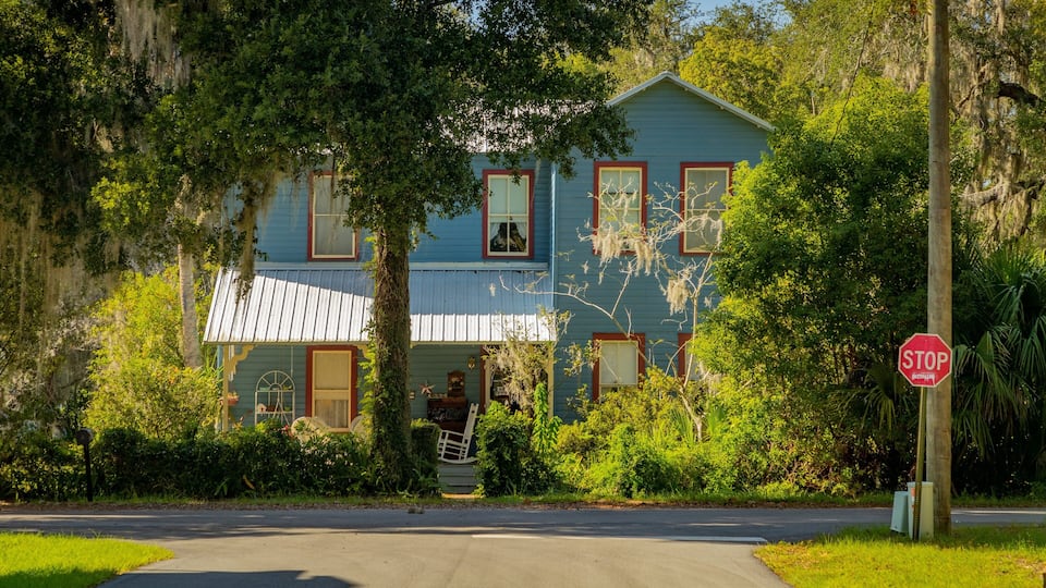 A blue house with red trim and a white roof, surrounded by lush greenery and a stop sign.