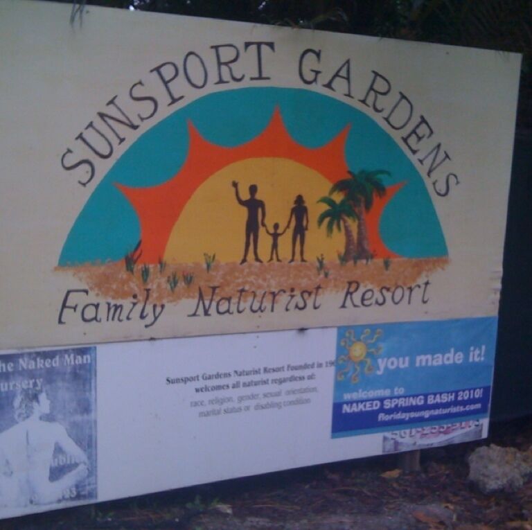 A sign for Sunport Gardens Family Naturist Resort with a sun and palm tree illustration.