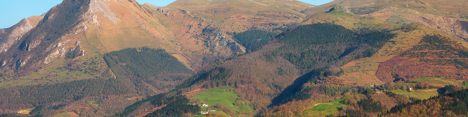 Un paisaje montañoso con un pico destacado, campos verdes y casas dispersas.