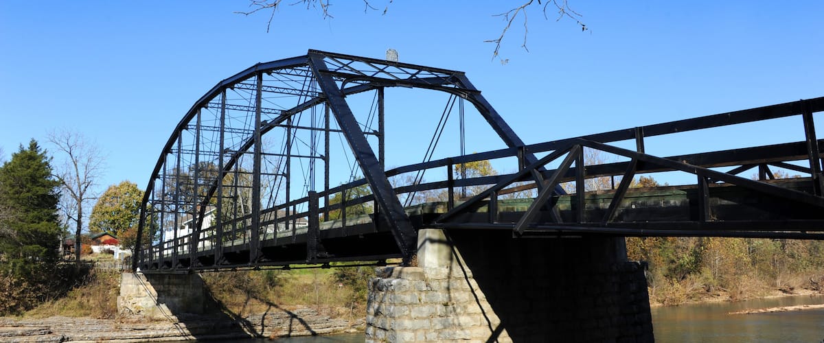 A steel bridge over a river with stone pillars, surrounded by trees and a clear blue sky.