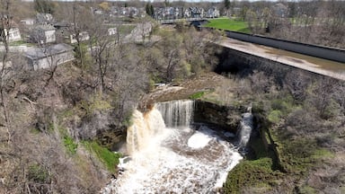 Ein Wasserfall stürzt über eine felsige Klippe, im Hintergrund sind eine Brücke und ein Wohngebiet zu sehen.