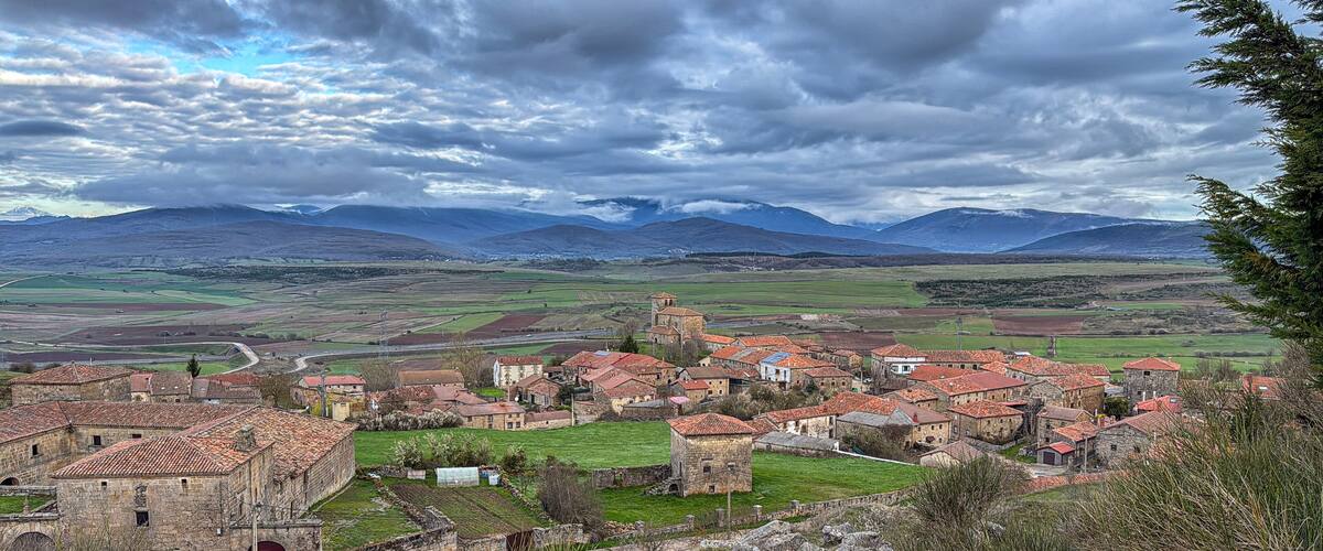 A historic village with red-tiled roofs, surrounded by green fields and mountains in the background.