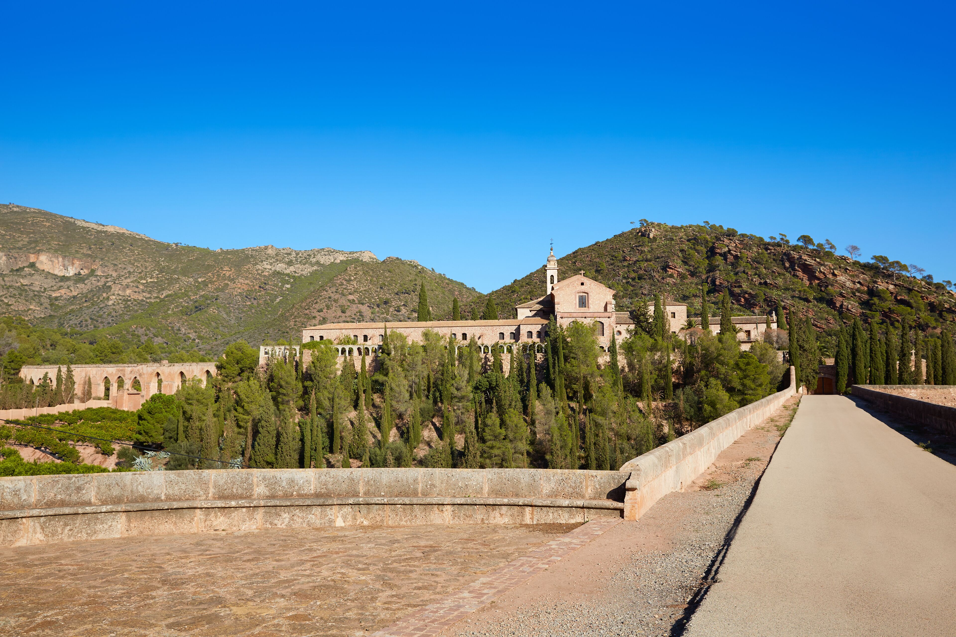A historic church with a bell tower, surrounded by a forested hillside and a clear blue sky.