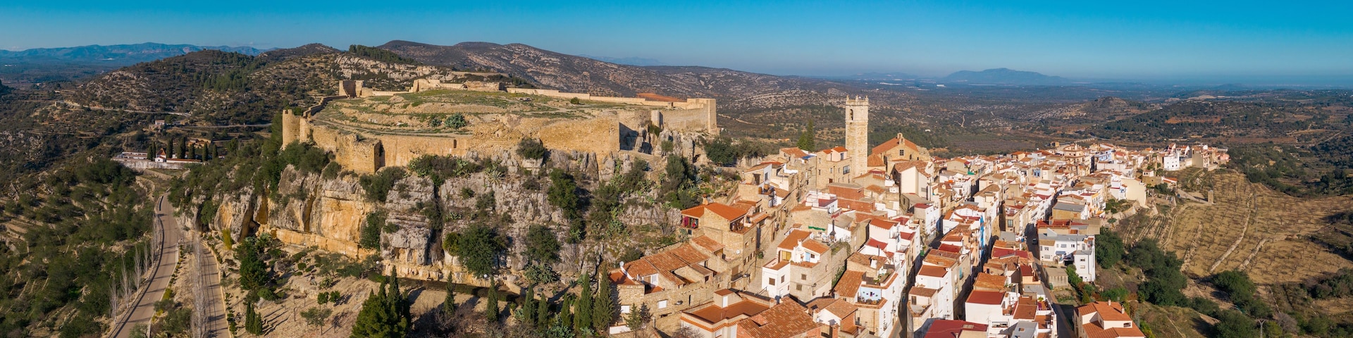 Un pueblo costero con un castillo imponente en una colina, viñedos en terrazas y un conjunto denso de edificios con tejados de tejas rojas.