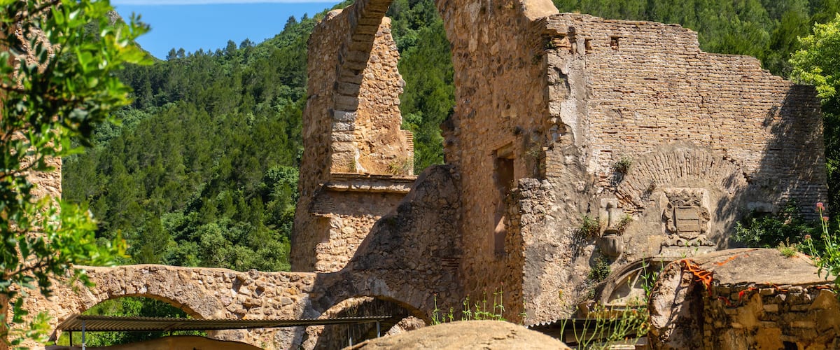 Ruínas antigas de pedra com um grande arco, rodeadas por vegetação exuberante e uma encosta arborizada.