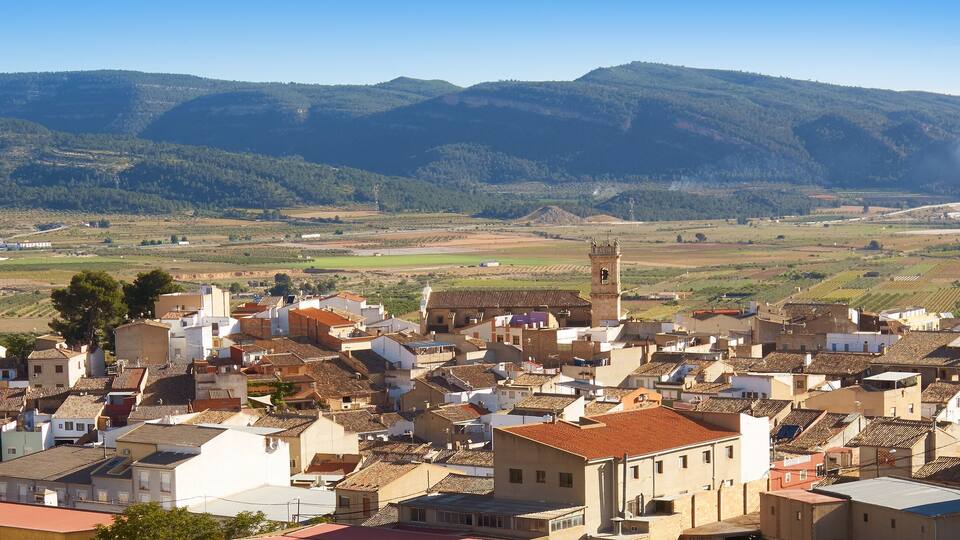 A town with terracotta-roofed buildings, a prominent church tower, and rolling hills in the background.