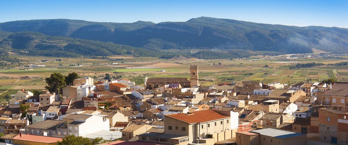A town with terracotta-roofed buildings, a prominent church tower, and rolling hills in the background.