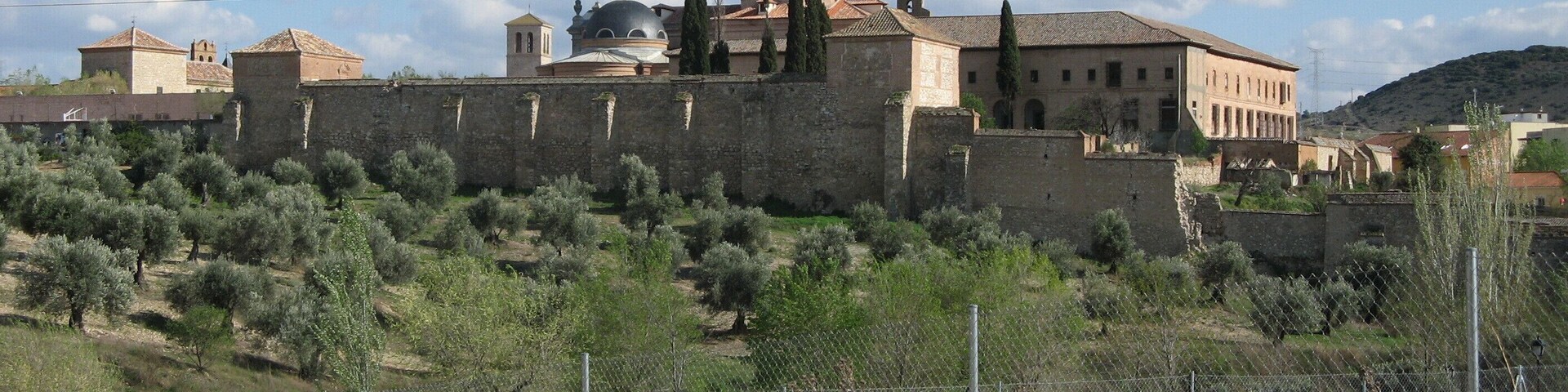 A historic building with a dome and bell tower, surrounded by a stone wall and olive trees.