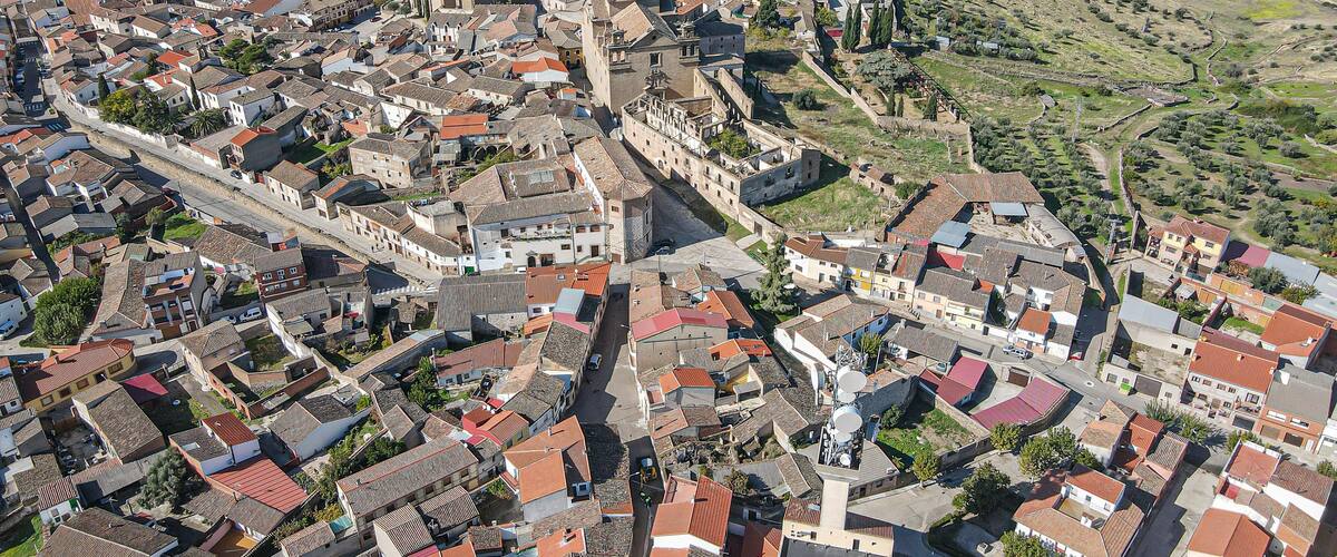 An aerial view of a historic town with a prominent castle, narrow streets, and surrounding fields.