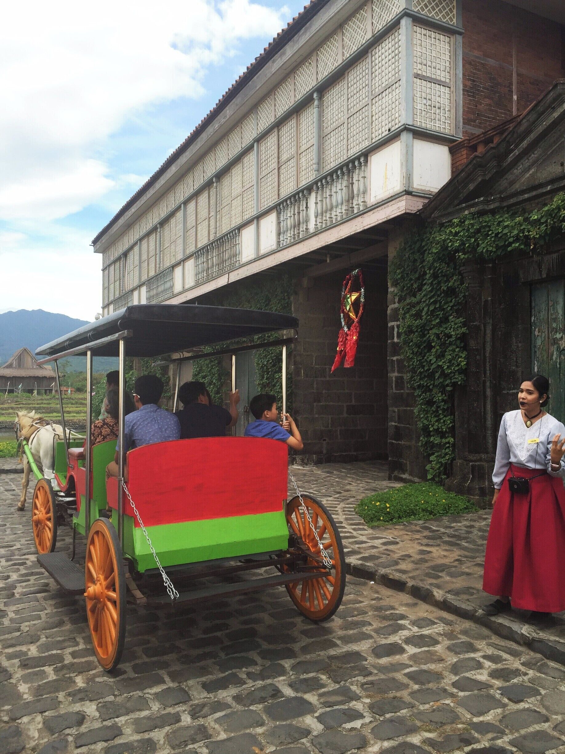 A horse-drawn carriage with passengers, a traditional building with lattice windows, and a woman in a skirt standing nearby.