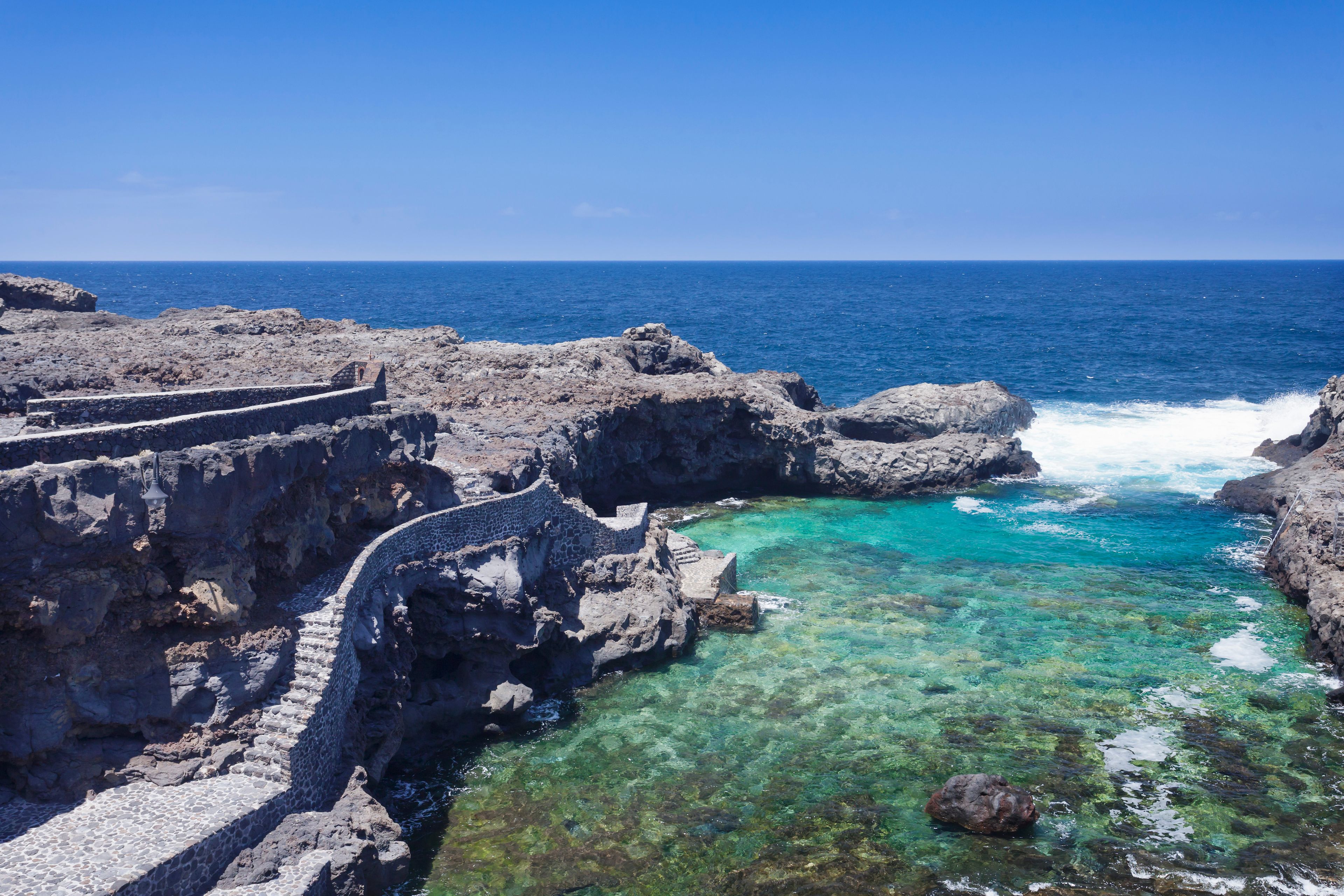 Eine Küstenlandschaft mit schroffen Klippen, klarem türkisblauem Wasser und einer Steintreppe, die zum Meer hinabführt.