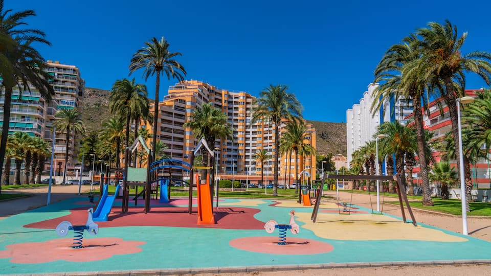 A playground with swings and slides, surrounded by palm trees and residential buildings.