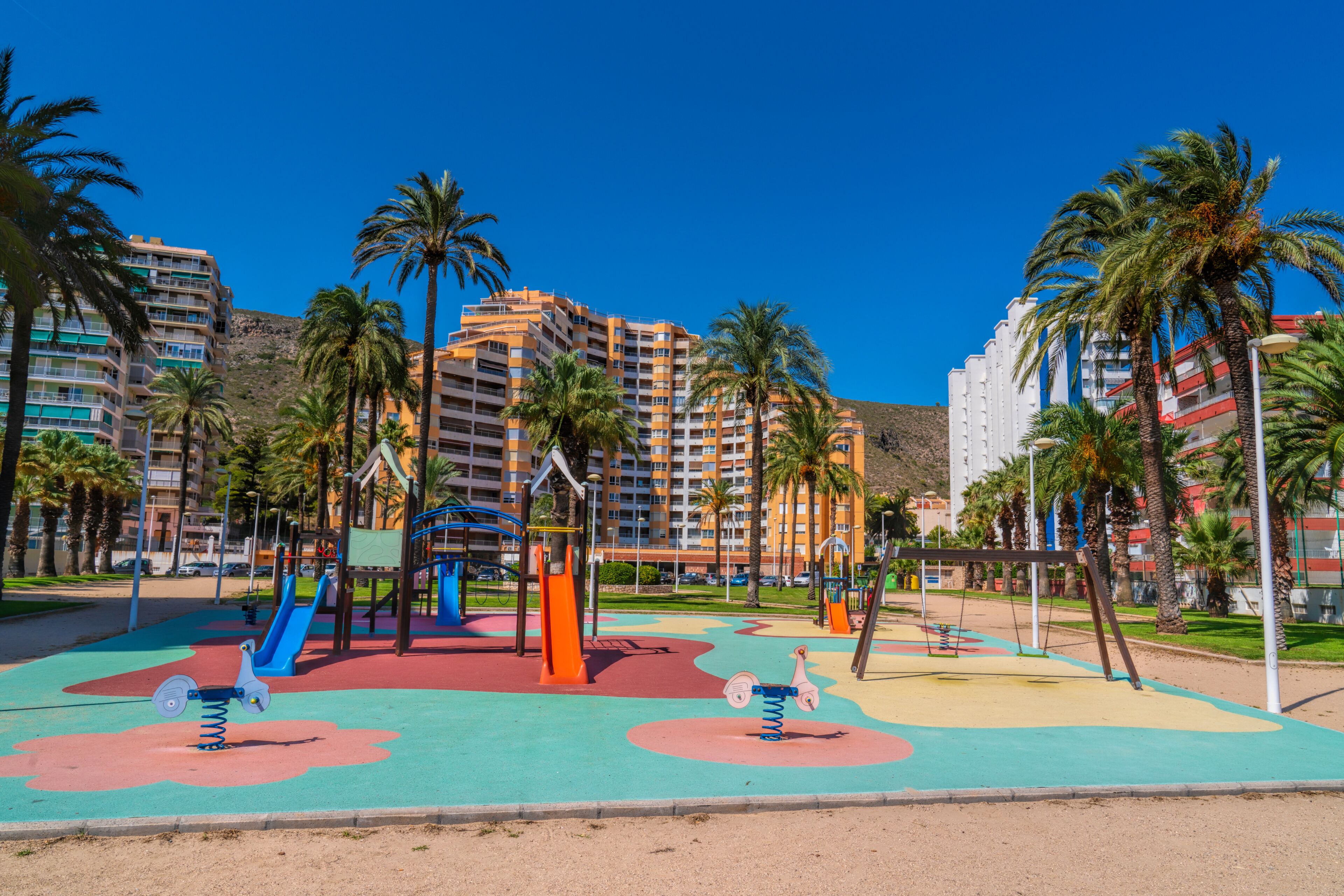 A playground with swings and slides, surrounded by palm trees and residential buildings.