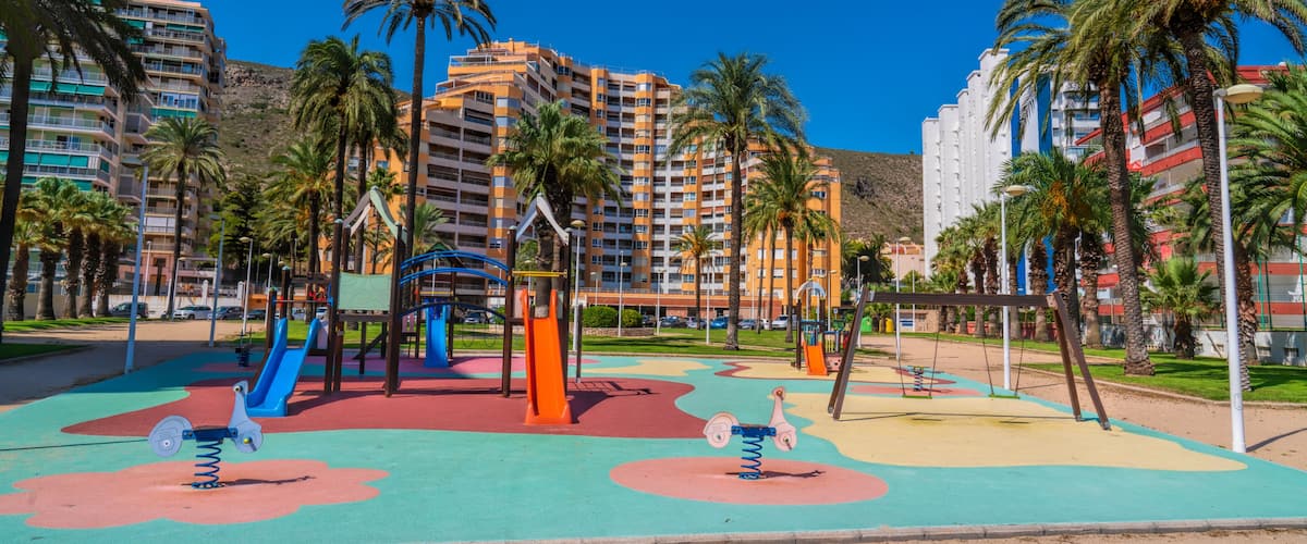 A playground with swings and slides, surrounded by palm trees and residential buildings.