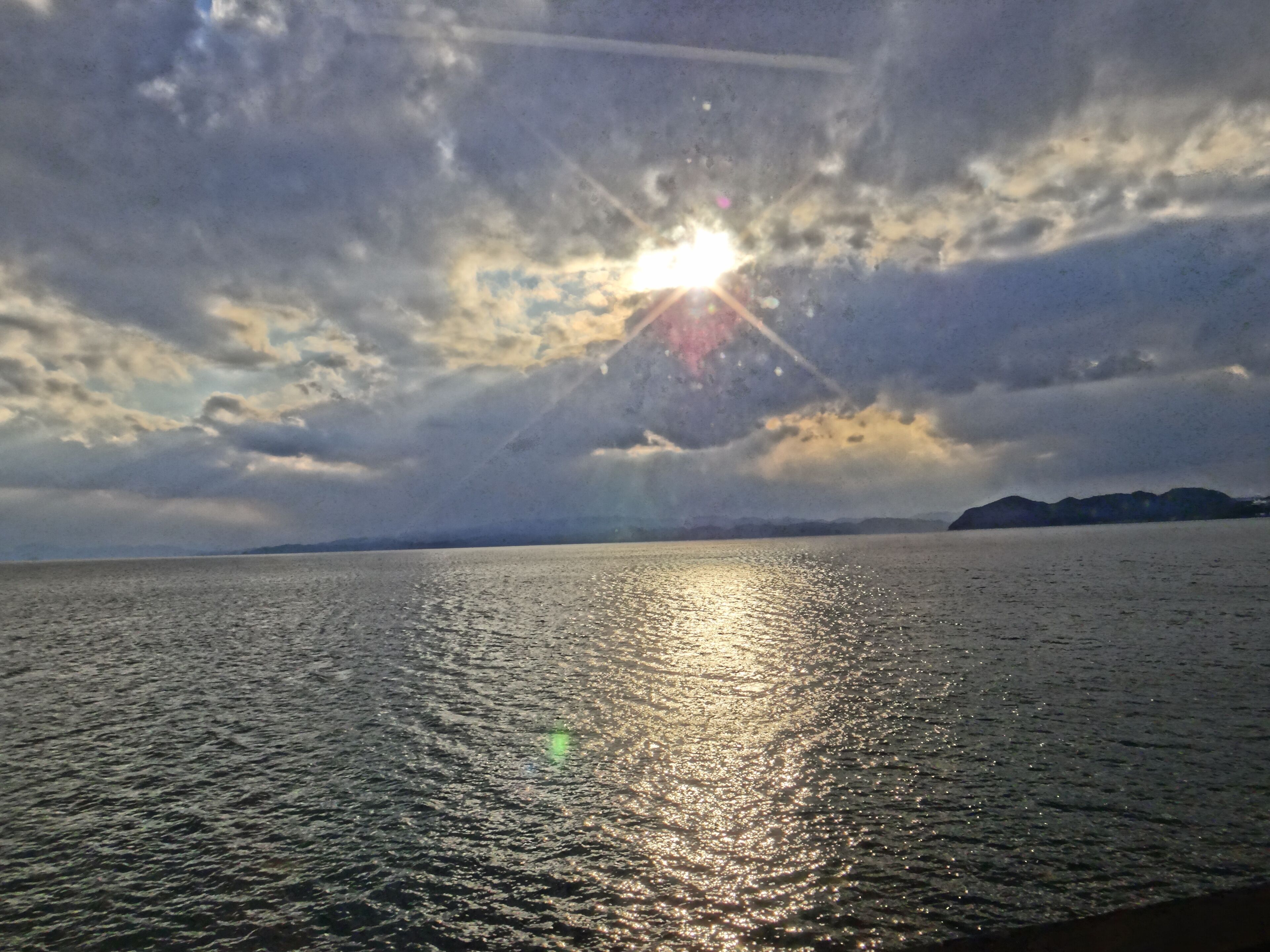 A body of water with sunlight reflecting off the surface, surrounded by a cloudy sky and distant landforms.