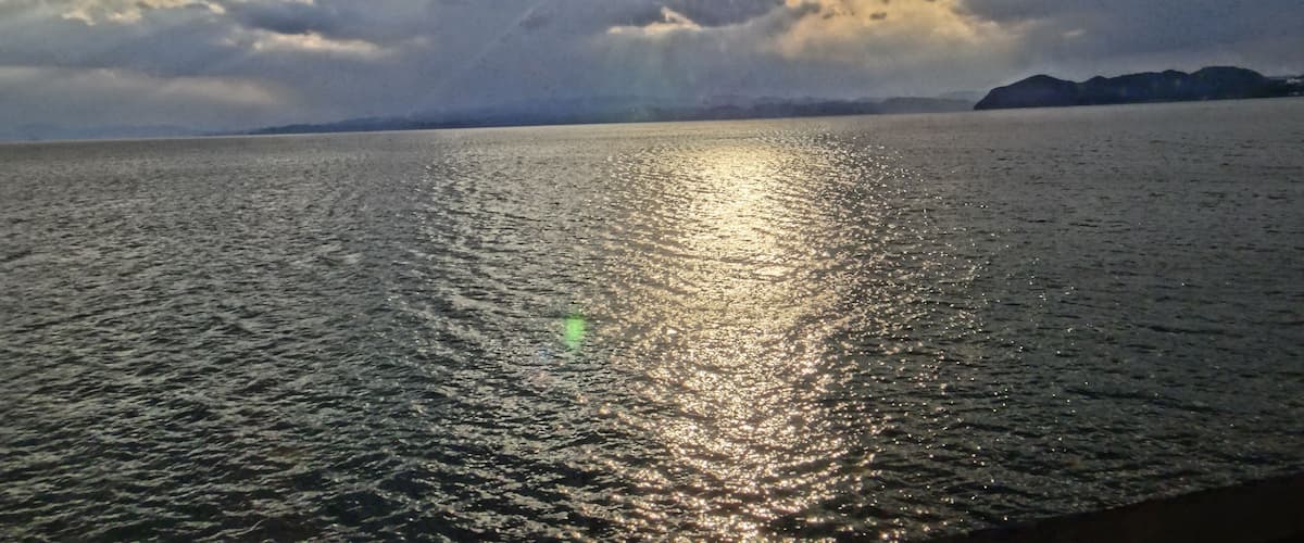 A body of water with sunlight reflecting off the surface, surrounded by a cloudy sky and distant landforms.
