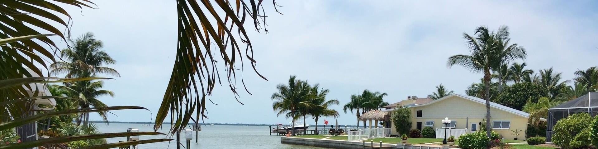 A wooden sign with 'Pine Island' and 'I live where you vacation' written on it, overlooking a body of water with palm trees and houses in the background.