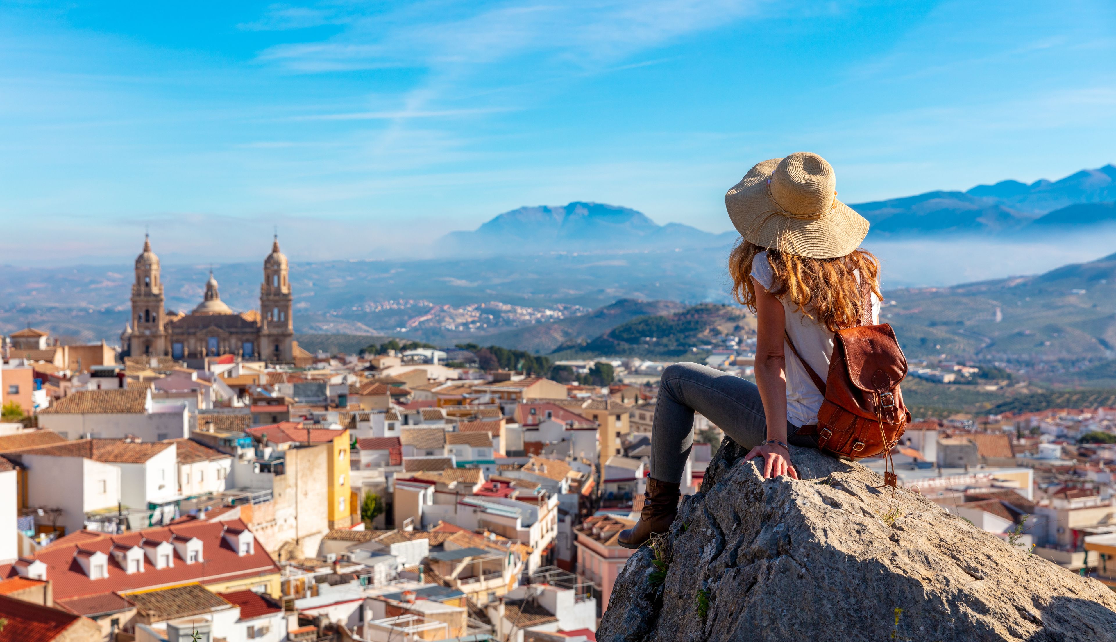Una persona sentada sobre una roca contemplando un paisaje urbano con edificios históricos y montañas al fondo.