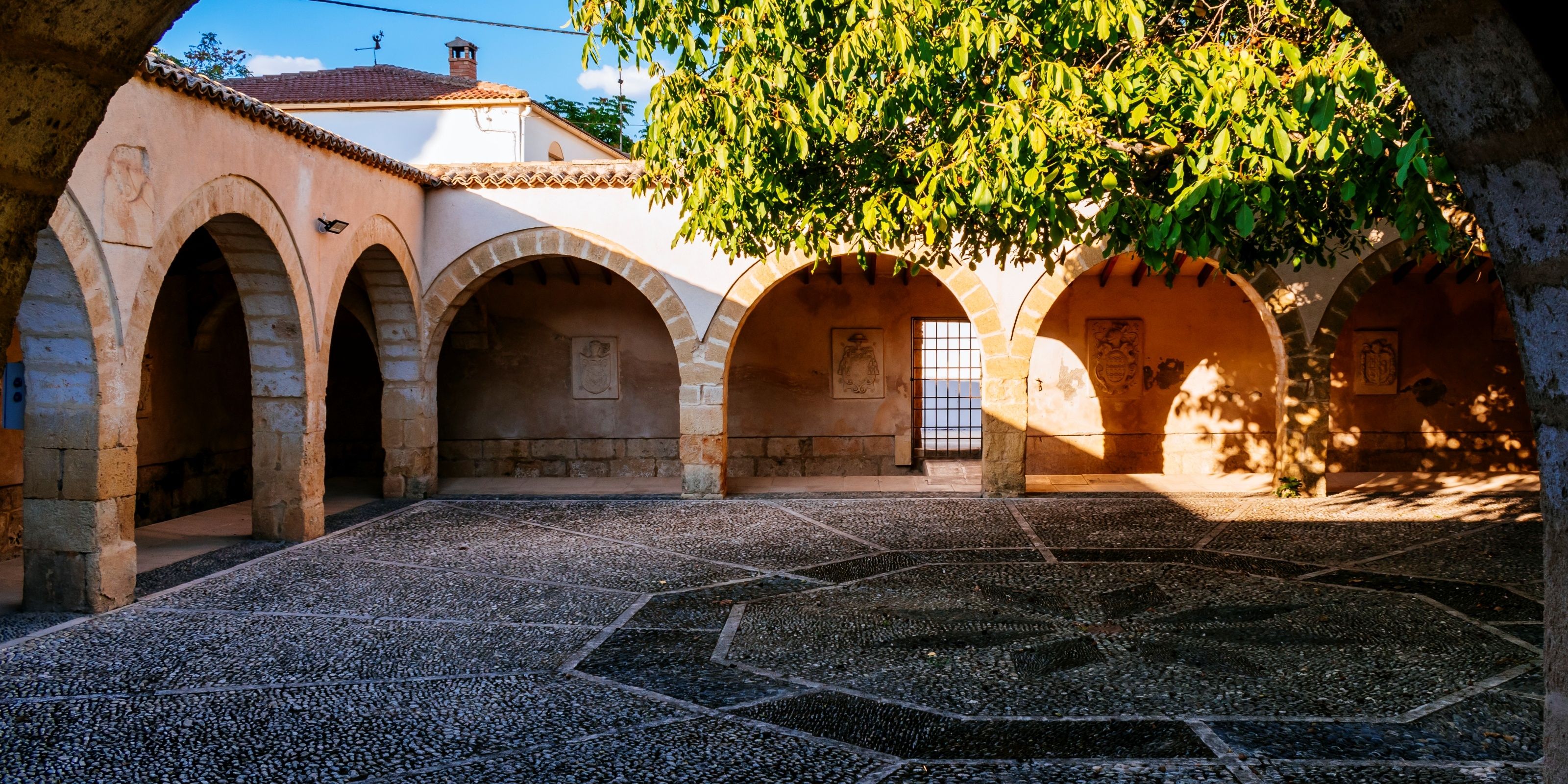 Un patio con arcos de piedra, suelo de baldosas y un árbol que da sombra.