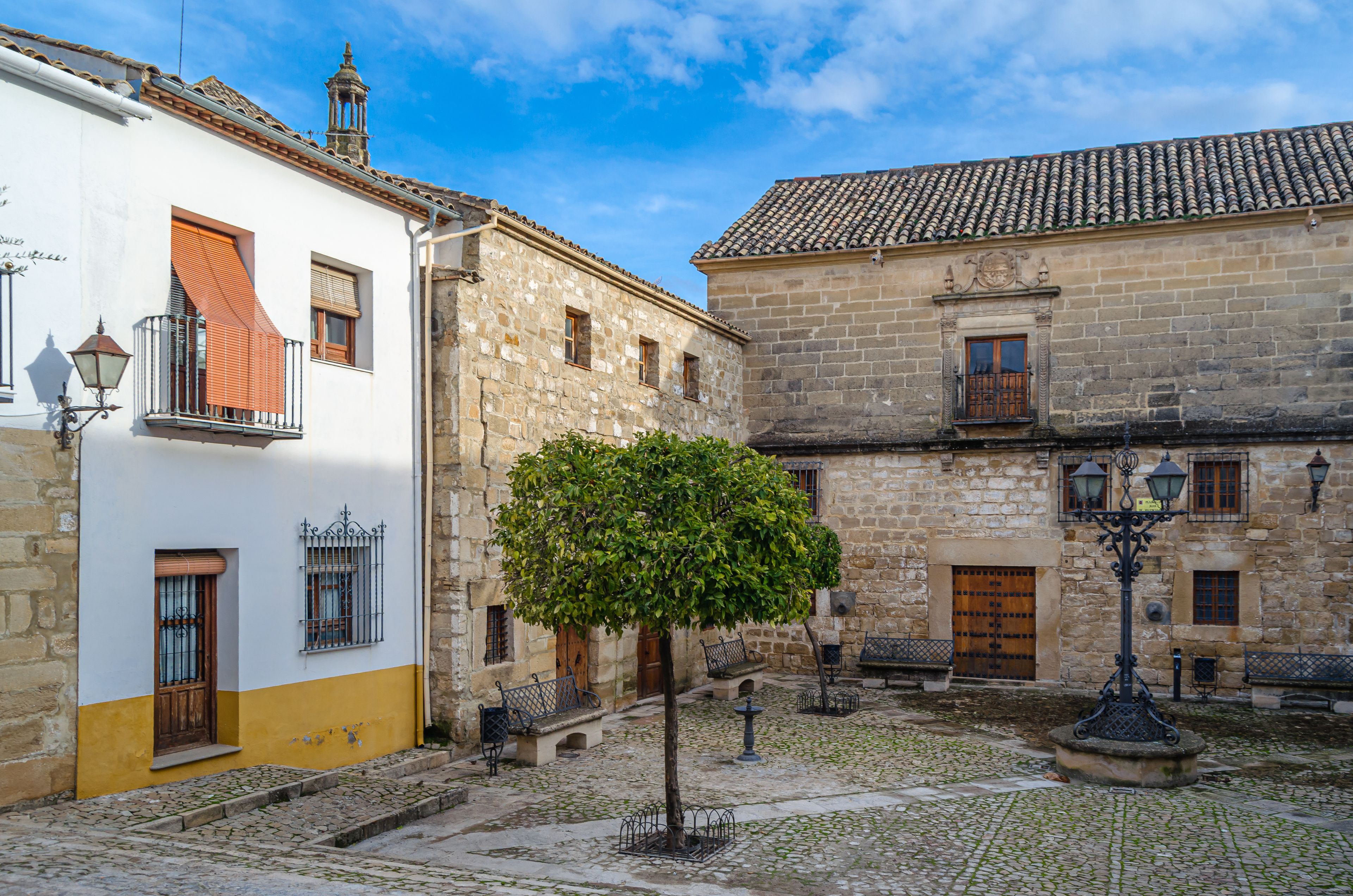 A cobblestone courtyard with historic stone buildings, a tree, and traditional street lamps.