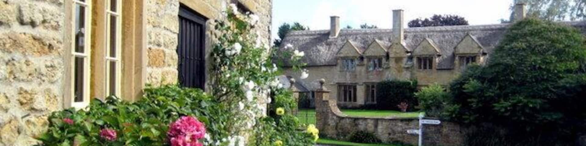A stone building with pink and white flowers in front, a paved road, and traditional houses in the background.