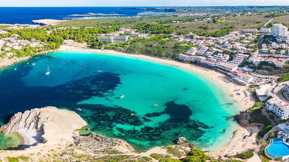A coastal town with a beach, clear turquoise waters, and a mountainous backdrop.