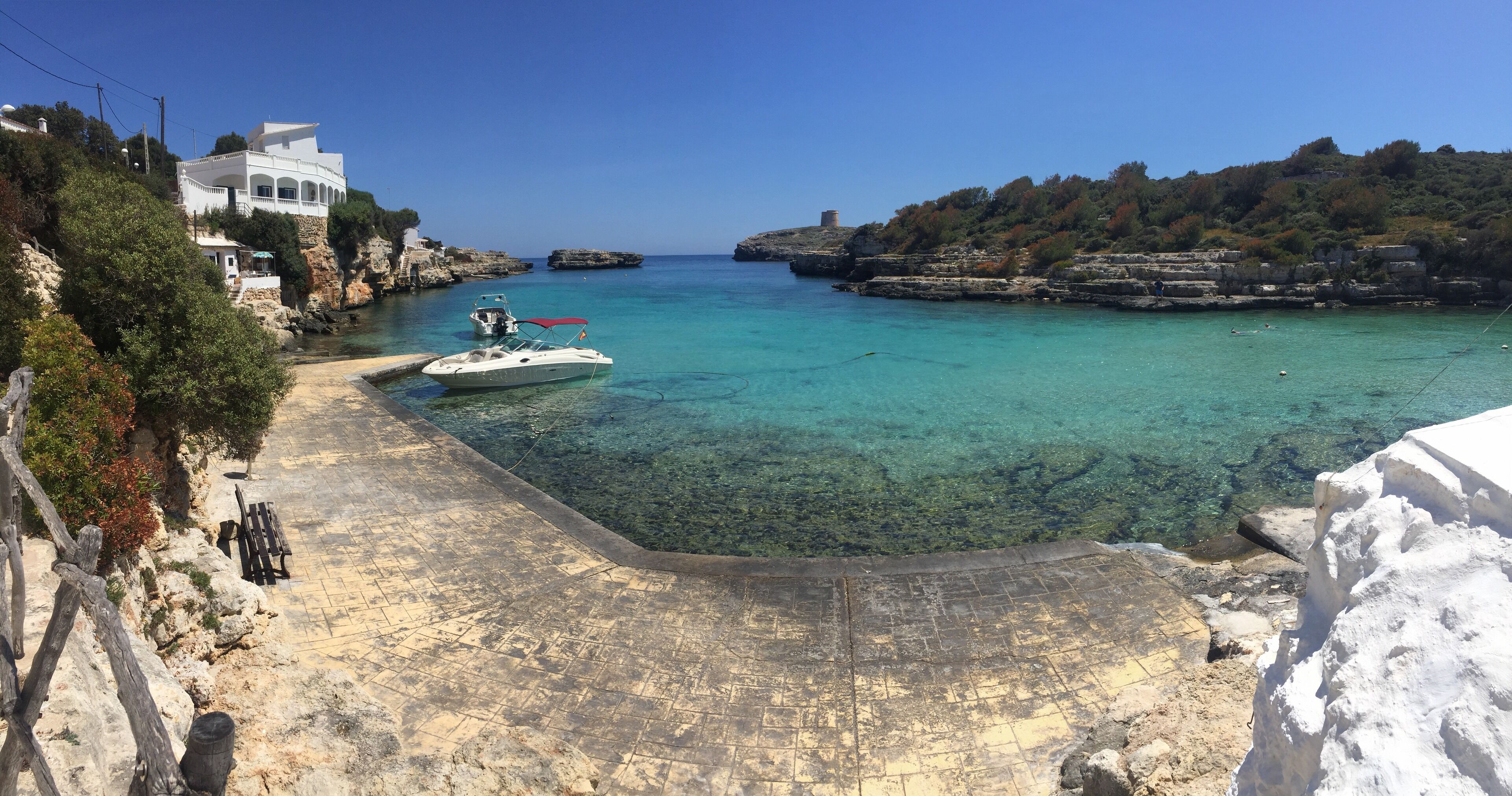A coastal scene with a clear blue sea, a white boat docked near a rocky shore, and a white building on a hillside.