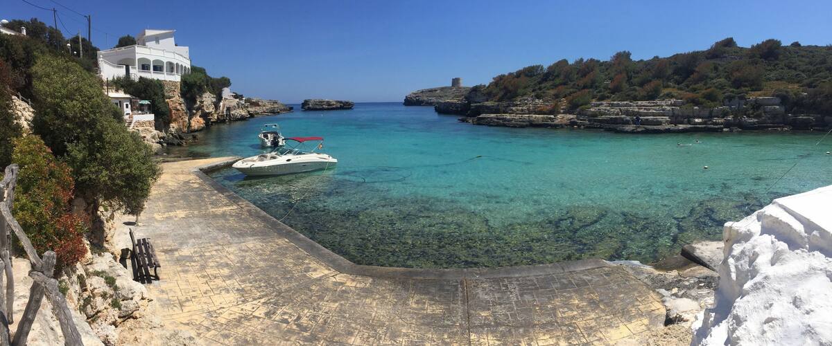 A coastal scene with a clear blue sea, a white boat docked near a rocky shore, and a white building on a hillside.