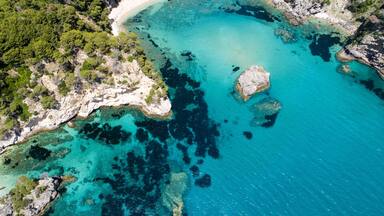 A coastal landscape with a sandy beach, clear turquoise waters, rocky outcrops, and dense green vegetation.