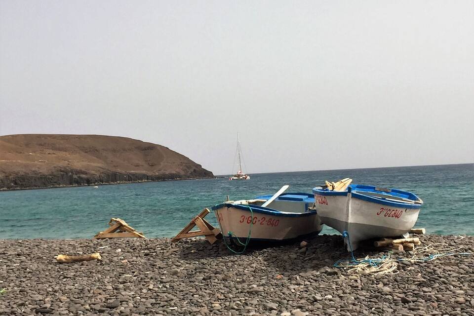 Twee boten op een kiezelstrand, met een zeilboot in de verte en een glooiend landschap.