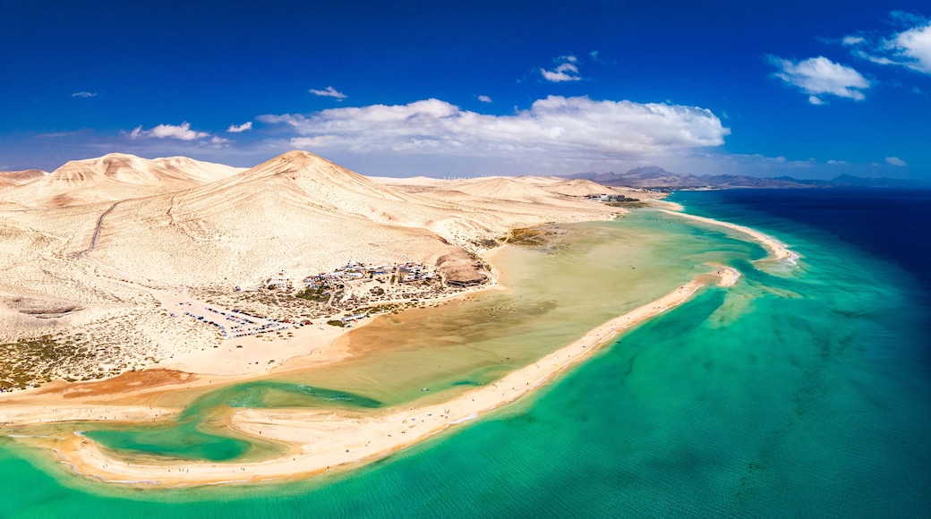 An aerial view of a coastal landscape with a prominent mountain range, a beach with clear turquoise waters, and a small settlement with buildings and roads.