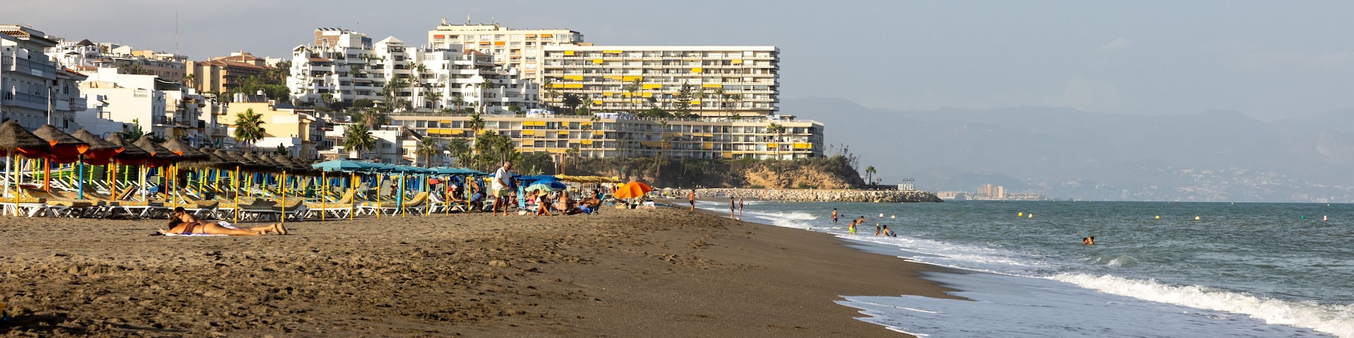 Uma praia com uma fileira de guarda-sóis coloridos e um grande edifício de hotel ao fundo.