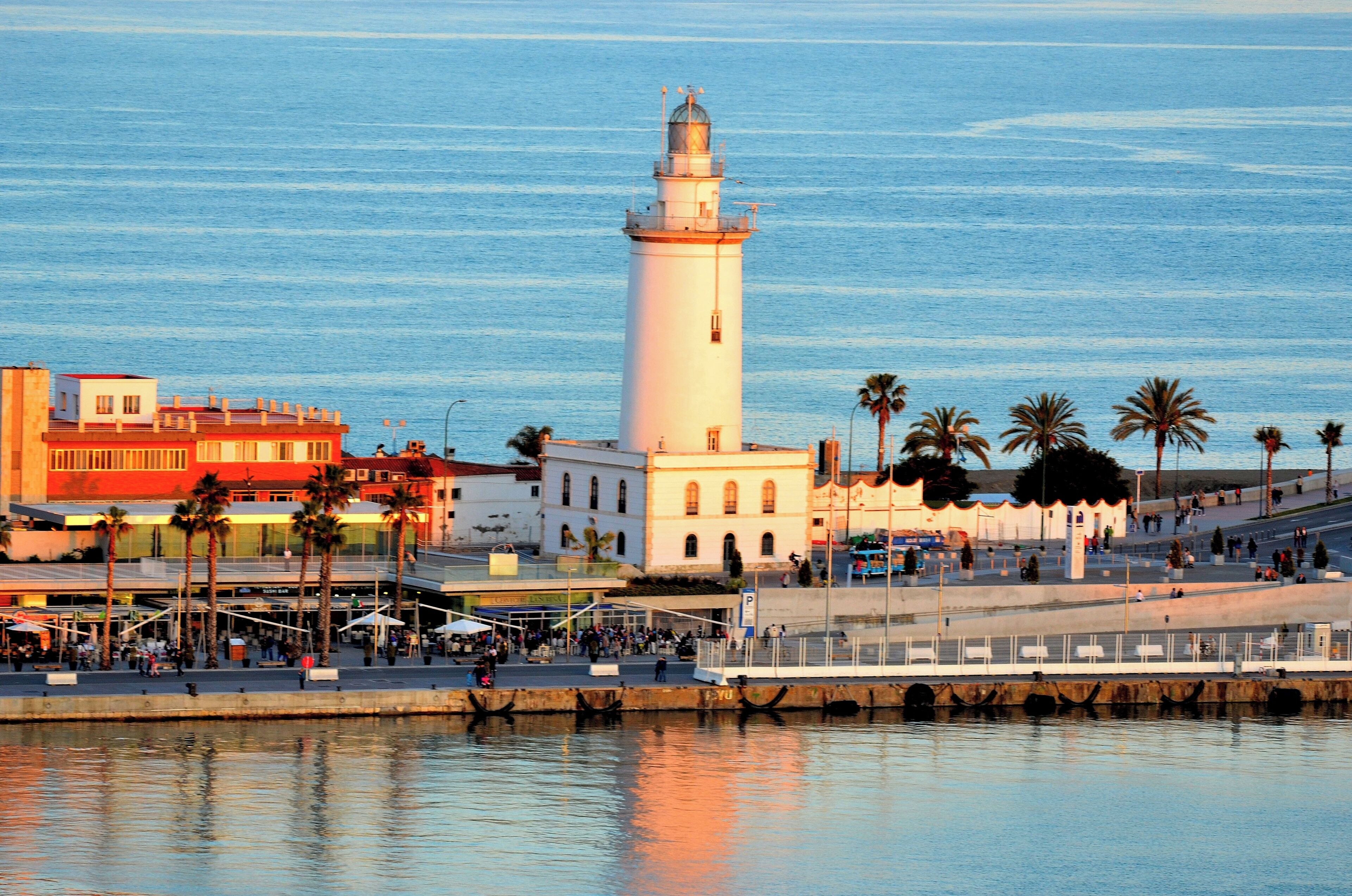 Un pueblo costero con un faro blanco destacado, palmeras y un paseo marítimo.