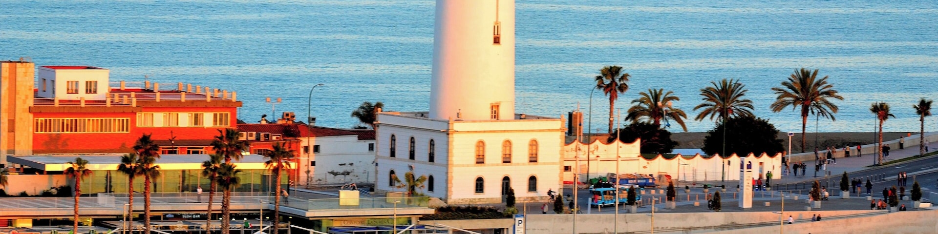 Un pueblo costero con un faro blanco destacado, palmeras y un paseo marítimo.