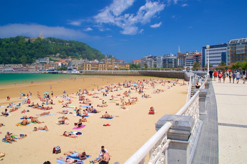 A crowded beach with people sunbathing and swimming, a clear blue sky, and buildings in the background.
