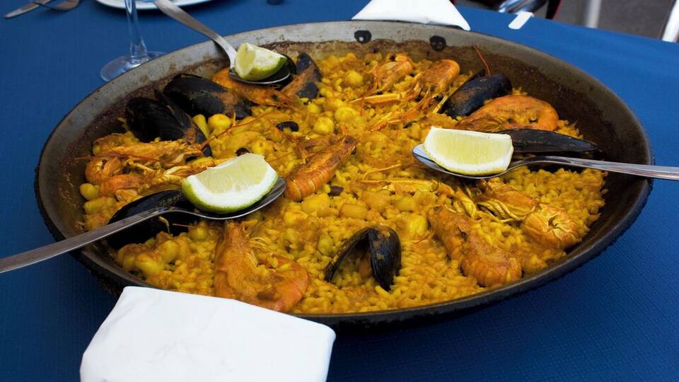 A large seafood paella with shrimp, mussels, and lemon wedges on a blue tablecloth.