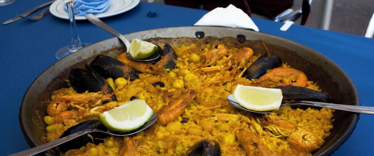 A large seafood paella with shrimp, mussels, and lemon wedges on a blue tablecloth.