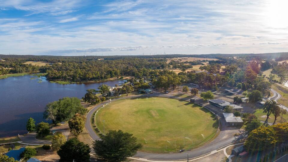 A lake surrounded by trees, a grassy field, and a road with buildings in the background.