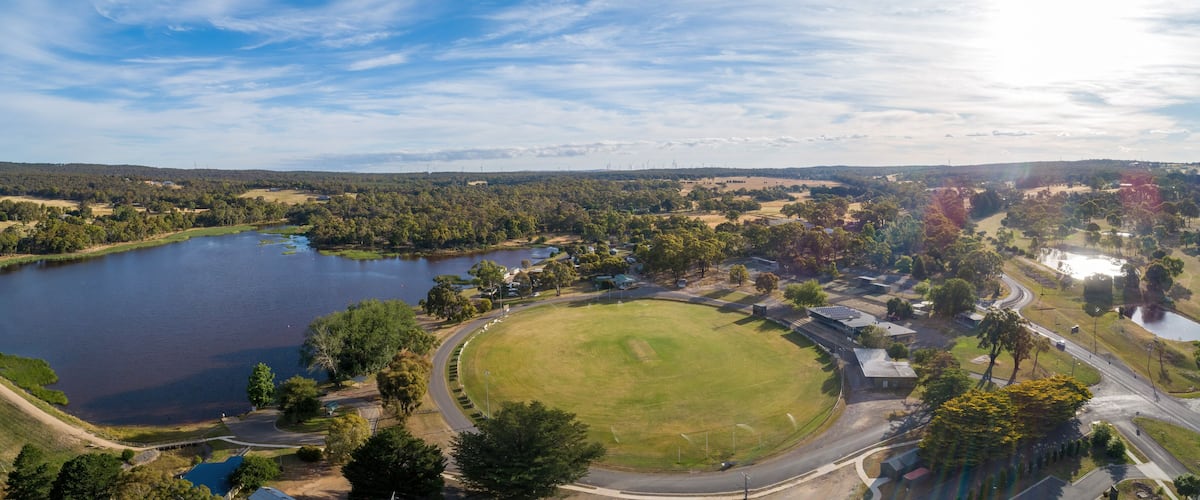 A lake surrounded by trees, a grassy field, and a road with buildings in the background.
