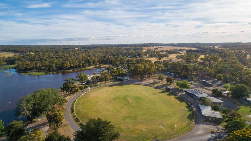 An aerial view of a park with a large grassy field, a lake, and surrounding trees.