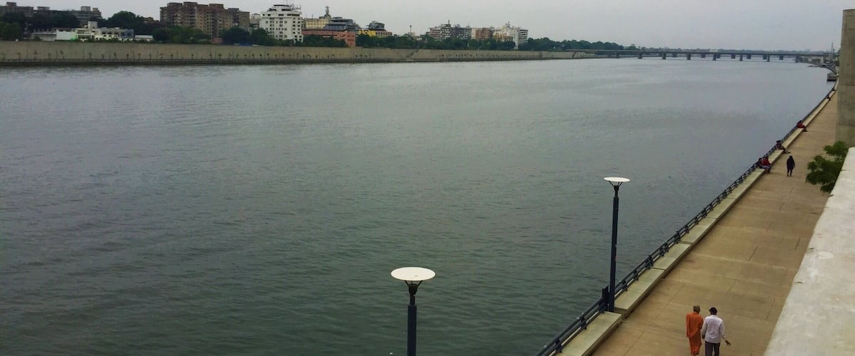 A river with a walkway and lamp posts, buildings in the background, and a bridge in the distance.