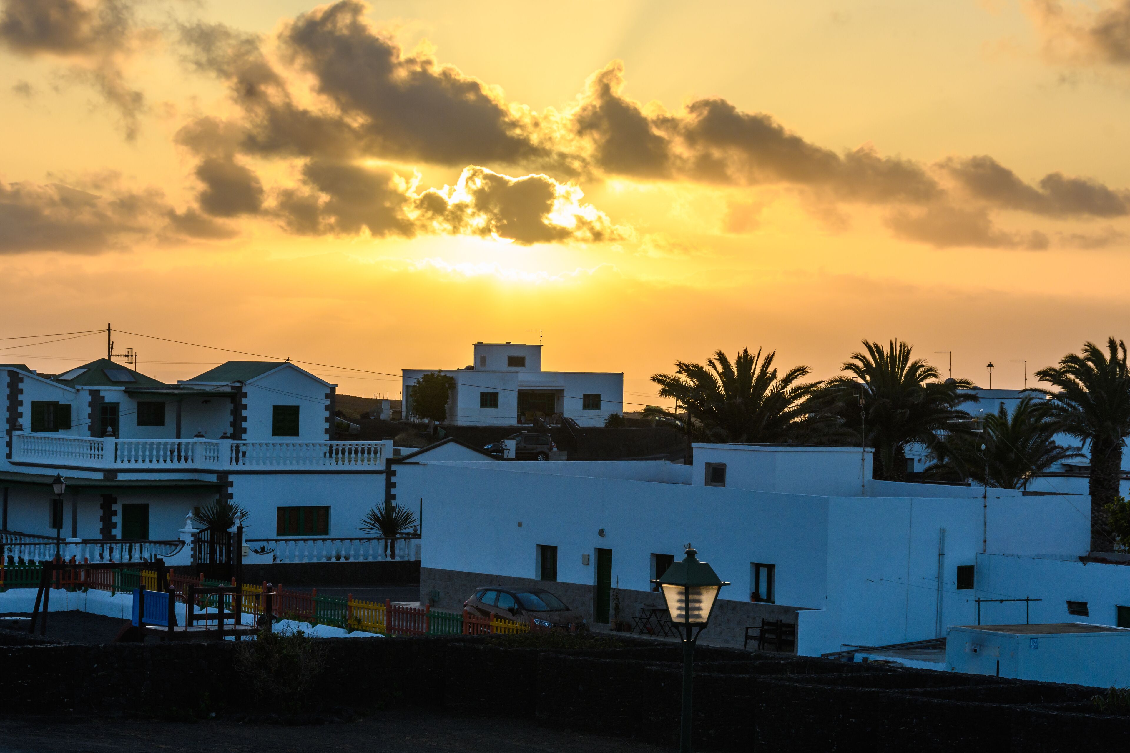 A sunset over a coastal town with white buildings, palm trees, and a street lamp.