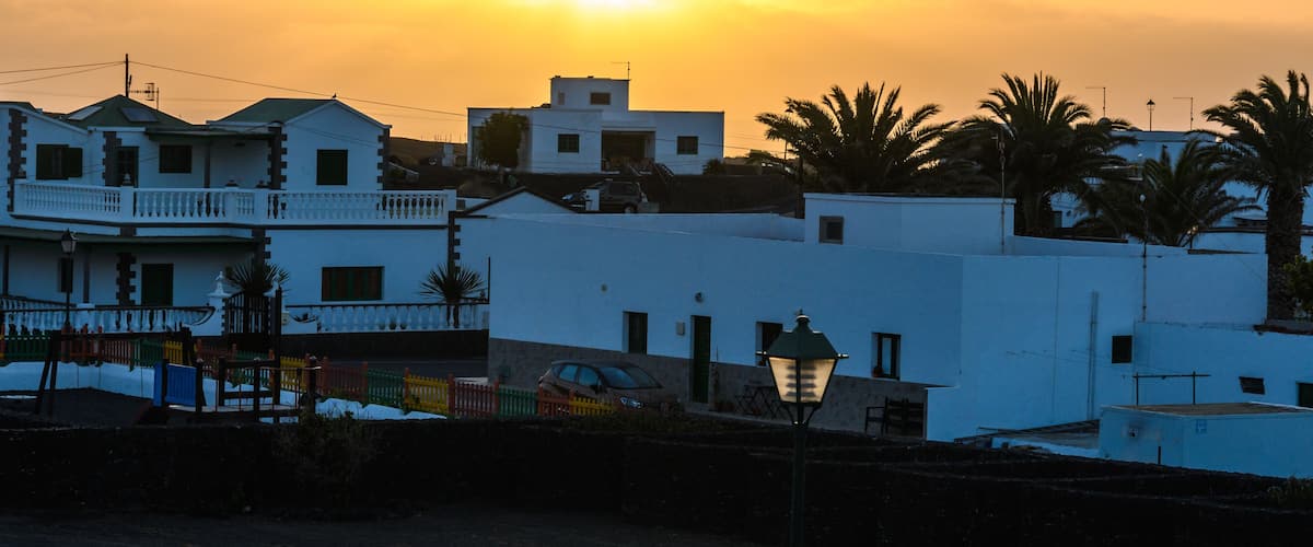 A sunset over a coastal town with white buildings, palm trees, and a street lamp.