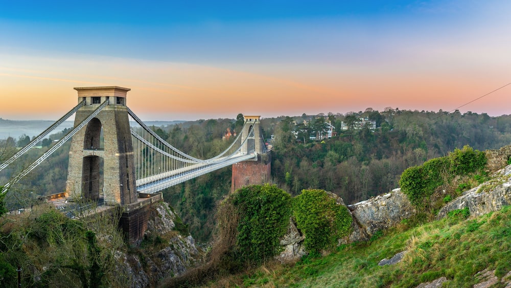 A suspension bridge with stone towers, surrounded by forested hills and a clear sky.