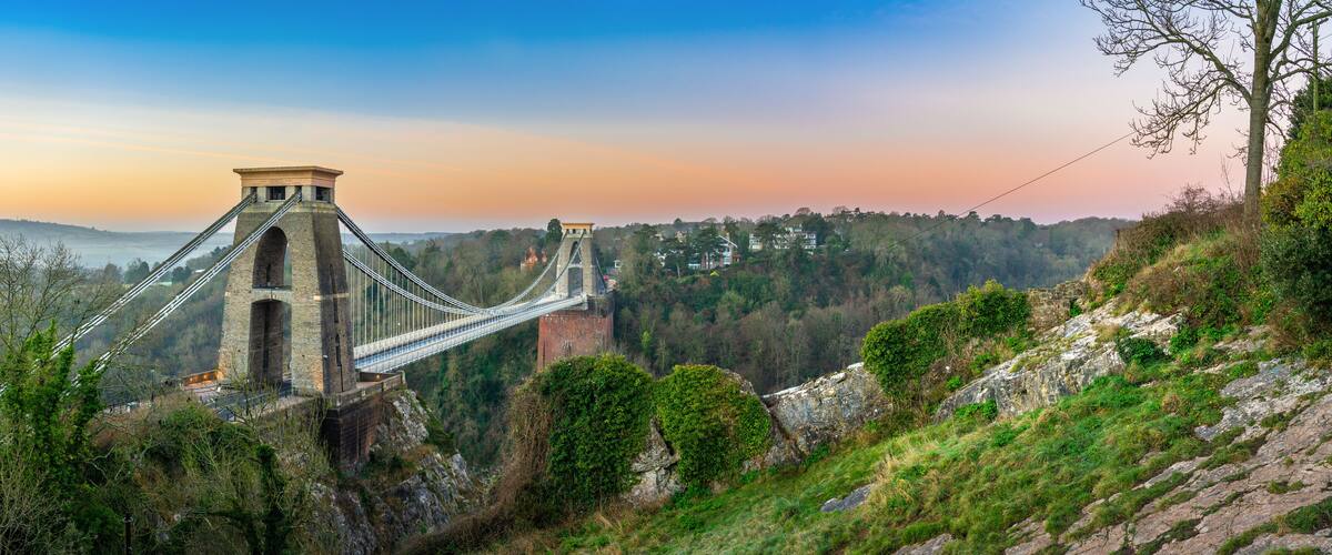 A suspension bridge with stone towers, surrounded by forested hills and a clear sky.